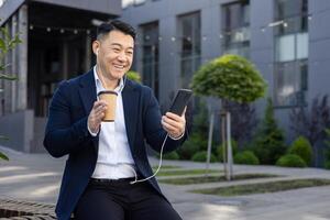 asiatico sorridente giovane uomo riposo su opera rompere, seduta su panchina vicino ufficio indossare cuffia, Tenere tazza di caffè e Telefono, parlando su chiamata. foto