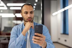 un' casualmente vestito uomo impegna con il suo smartphone, navigazione o comunicare, mentre in piedi nel un' contemporaneo ufficio ambiente con un' fiducioso postura. foto