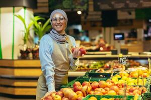 ritratto di commessa nel supermercato, contento donna nel hijab sorridente e guardare a telecamera, venditore Tenere mele nel verdura sezione, musulmano donna nel bicchieri e grembiule tra scaffali con merce foto