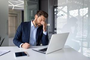 stressato uomo d'affari con mano su fronte seduta a scrivania con computer portatile, tavoletta, e lavoro d'ufficio nel moderno ufficio. foto