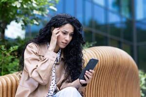 pensiero e irritato donna al di fuori ufficio edificio con Telefono, ispanico attività commerciale donna frustrato lettura notizia in linea e navigazione Internet pagine, utilizzando smartphone. foto