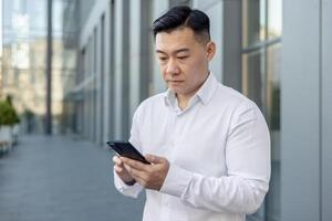 un' grave giovane asiatico uomo nel un' bianca camicia è in piedi vicino un ufficio edificio al di fuori e utilizzando un' mobile Telefono nel concentrazione. foto