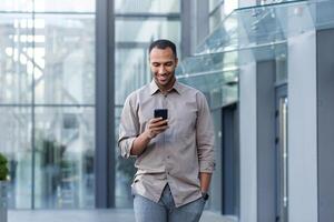 latino americano uomo d'affari nel casuale camicia a piedi vicino ufficio edificio a partire dal fuori, uomo sorridente e utilizzando Telefono, libero professionista digitando Messaggio e navigazione Internet pagine foto