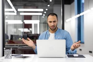 perplesso giovane uomo d'affari guardare a il suo il computer portatile schermo nel un' contemporaneo ufficio, mostrando un' senso di confusione e incertezza. foto