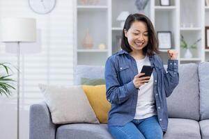 donna festeggiare vittoria a casa guardare a Telefono schermo, soddisfatto asiatico donna contento ricevuto in linea notifica di bene realizzazione risultati seduta su divano nel vivente camera. foto