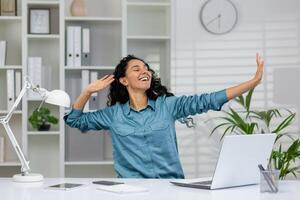 esuberante giovane donna con braccia sollevato nel trionfo, seduta a sua casa ufficio con un' il computer portatile e un' allegro Sorridi. foto