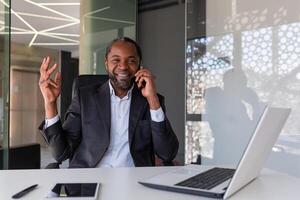 ritratto di riuscito africano americano uomo dentro ufficio a posto di lavoro, maturo uomo d'affari parlando su Telefono sorridente e guardare a telecamera, soddisfatto con finanziario realizzazione capo con il computer portatile. foto