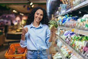 ritratto di un' bellissimo vegetariano donna nel un' supermercato, un' ispanico donna sorridente e guardare a il telecamera e sorridente sceglie broccoli verdure nel un' drogheria memorizzare foto