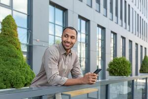 allegro giovane alunno nel camicia utilizzando Telefono al di fuori Università città universitaria, africano americano uomo sorridente e guardare lontano, Tenere smartphone, digitando Messaggio e fabbricazione chiamata. foto