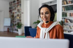 insegnante conduce Lezioni in linea, donna con chiamata cuffia sorridente e spiegando conferenza in linea utilizzando il computer portatile per a distanza comunicazione e apprendimento, seduta a scrivania nel Università biblioteca a scrivania. foto