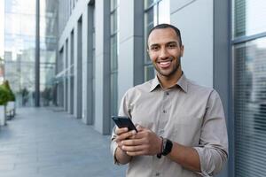 ritratto di africano americano uomo d'affari, uomo Tenere Telefono sorridente e guardare a telecamera, ufficio lavoratore nel camicia al di fuori moderno ufficio edificio su pranzo rompere foto