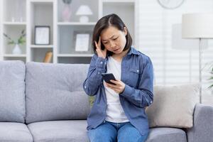 giovane asiatico donna irritato seduta a casa, donna avuto cattivo notizia in linea su Telefono seduta su divano nel vivente camera, irritato e deluso. foto