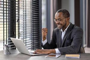 un adulto maschio uomo d'affari nel un' completo da uomo celebra successo mentre Lavorando su il suo il computer portatile a un' moderno ufficio spazio. foto