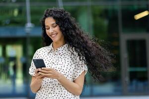 giovane bellissimo latino americano donna a piedi nel il città con Telefono nel mani, attività commerciale donna dopo Lavorando giorno, sorridente con soddisfazione, alunno utilizzando applicazione su smartphone. foto