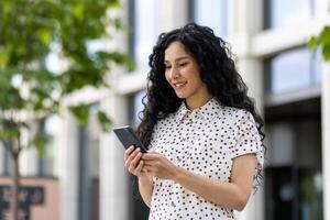 un' giovane bellissimo latino americano donna con Riccio capelli passeggiate attraverso il città con un' Telefono nel sua mani, un' donna usi un applicazione su un' smartphone, sfoglia Internet pagine e sociale reti. foto