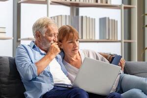 anziano coppia la spesa tempo insieme nel il vivente camera, donna e uomo utilizzando il computer portatile e digitale tavoletta su il accogliente divano a casa foto
