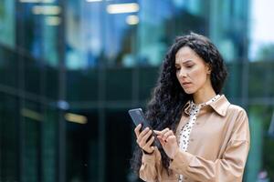 irritato e triste donna con Telefono nel mani lettura in linea Messaggio, ispanico attività commerciale donna ricevuto cattivo notizia notifica mentre a piedi al di fuori ufficio edificio con smartphone. foto