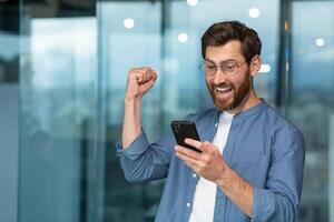 riuscito uomo d'affari Lavorando nel moderno ufficio costruzione, uomo guardare a smartphone e lettura notizia festeggiare contento notizia vittoria uomo d'affari nel camicia e bicchieri Tenere mano su trionfo gesto. foto