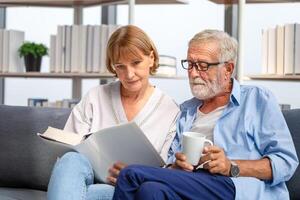 anziano coppia la spesa tempo insieme nel il vivente camera, donna e uomo lettura un' libro e godendo caffè su il accogliente divano a casa foto