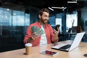 riuscito uomo d'affari nel rosso camicia festeggiare vittoria e bene realizzazione Tenere Telefono e i soldi americano denaro contante dollari nel mani, investitore Lavorando dentro ufficio con il computer portatile avuto reddito. foto