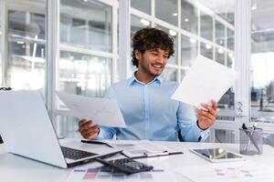 riuscito finanziere contabile Lavorando dentro ufficio con il computer portatile e documenti, uomo soddisfatto con revisione risultati sorridente e lettura finanziario rapporti, uomo d'affari utilizzando il computer portatile per scartoffie. foto