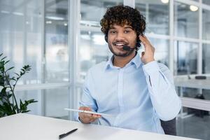 un' allegro maschio cliente servizio rappresentante con Riccio capelli indossare un' cuffia, sorridente e Lavorando nel un' moderno ufficio ambiente. foto