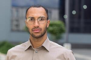 vicino su foto ritratto di giovane uomo d'affari uomo nel camicia, grave e riflessivo uomo guardare a telecamera indossare bicchieri al di fuori moderno ufficio edificio