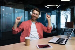 un' uomo d'affari nel un' rosso camicia e bicchieri ascolta per musica nel cuffia, un' uomo balli mentre seduta a il posto di lavoro riposo, il capo usi un' il computer portatile a opera. foto