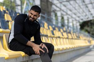 un' maschio atleta nel allenarsi abbigliamento sperimentare ginocchio dolore mentre seduta su giallo stadio sedili, evidenziazione gli sport infortunio e fitness concetti. foto