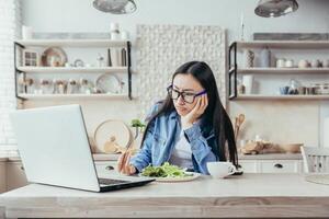 pranzo mentre Lavorando in linea. un' giovane asiatico donna si siede nel il cucina a casa con un' il computer portatile e insoddisfatto mangia il pranzo, fresco insalata, verdure. odia dieta, salutare stile di vita foto