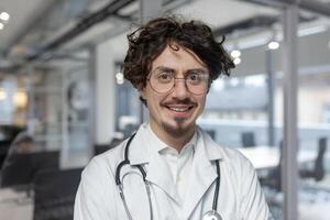 un' medico nel un' bianca medico cappotto sta nel il suo ufficio, Tenere un' stetoscopio. uomo vicino su guarda dentro telecamera Sorridi foto