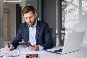 un' focalizzata maschio contabile analizzando lavoro d'ufficio mentre calcolatore spese utilizzando il computer portatile e calcolatrice a il suo ufficio scrivania. foto