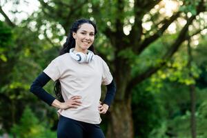 orgoglioso misto donna con lungo Riccio capelli e sportivo uniforme in piedi all'aperto con mani su vita. soddisfatto giovane signora completato quotidiano allenarsi e sensazione motivata per raggiungimento più grande risultati. foto