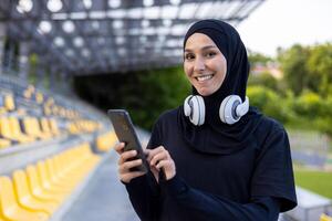 ritratto di un' giovane hijab musulmano donna nel un' gli sport stadio in esecuzione e fare attivo fisico esercizi, sorridente e guardare a il telecamera, utilizzando il Telefono e cuffie per ascolta per musica. foto