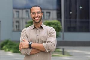 ritratto di giovane ispanico alunno uomo sorridente e guardare a telecamera vicino Università città universitaria con braccia attraversato indossare bicchieri e camicia foto