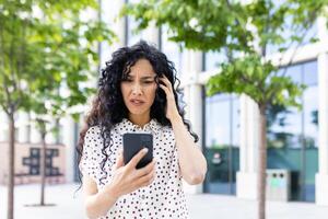 ansioso giovane donna con Riccio capelli guardare a Telefono schermo con un' ha riguardato espressione, in piedi all'aperto vicino ufficio edifici. foto