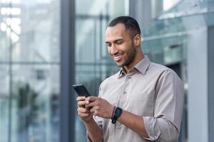 ispanico uomo al di fuori moderno ufficio edificio utilizzando smartphone, uomo d'affari nel camicia digitando Messaggio e navigazione in linea pagine foto