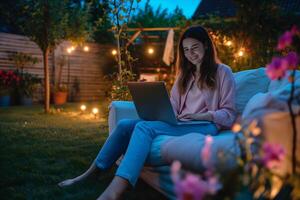 donna sorridente si siede su un' confortevole divano guardare a il il computer portatile a notte nel il Giardino dietro la casa foto