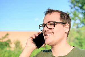 giovane uomo con occhiali e verde maglietta parlando su cellula Telefono a il parco foto