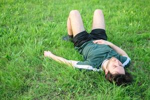 giovane uomo con verde maglietta dire bugie su erba nel parco dopo esercizio esausto foto