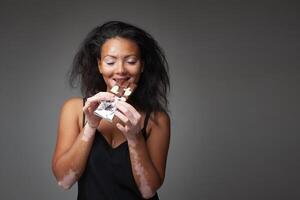 sorridente nero donna con vitiligine malattia mangiare cioccolato nel grigio studio foto