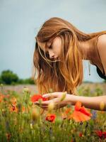 un' bellissimo giovane ragazza nel un' nero sera toga piegato al di sopra di per odore il fragranza di un' fiore nel un' papavero campo su un' nuvoloso estate giorno. ritratto di un' femmina modello all'aperto. piovoso tempo atmosferico. grigio nuvole. foto