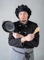 giovane chef in uniforme nera che tiene una casseruola e un cucchiaio isolati su sfondo grigio foto