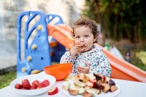 poco ragazza mangia mentre in piedi nel davanti di un' impostato tavolo con tagliare frutta su piatti nel il giardino foto