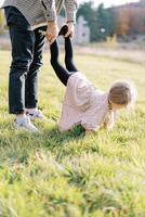 papà detiene il gambe di un' poco ragazza a piedi su sua mani lungo il verde prato. ritagliata foto