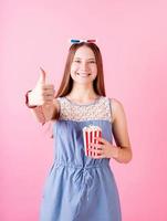 Sorridente ragazza adolescente che indossa occhiali 3D mangiare popcorn isolato su sfondo rosa che mostra il pollice in alto foto