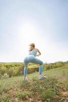 un atletico ragazza esegue squat per esercizio sua gluteo coscia muscoli. bellissimo bionda caucasico donna nel blu stretto tuta. bionda ragazza a un all'aperto formazione sessione foto