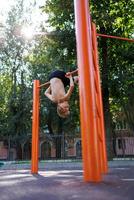 un' adolescente nel il parco va nel per gli sport sospeso su il bar a il parte inferiore con il suo testa. strada allenarsi su un' orizzontale bar nel il scuola parco. foto
