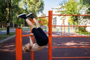 un' adolescente esegue esercizi di lancio il suo gambe al di sopra di il orizzontale sbarra. strada allenarsi su un' orizzontale bar nel il scuola parco. foto
