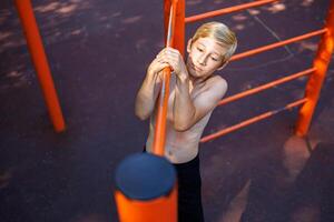 gli sport bambino esegue un' lato pull-up su il traversa. strada allenarsi su un' orizzontale bar nel il scuola parco. foto
